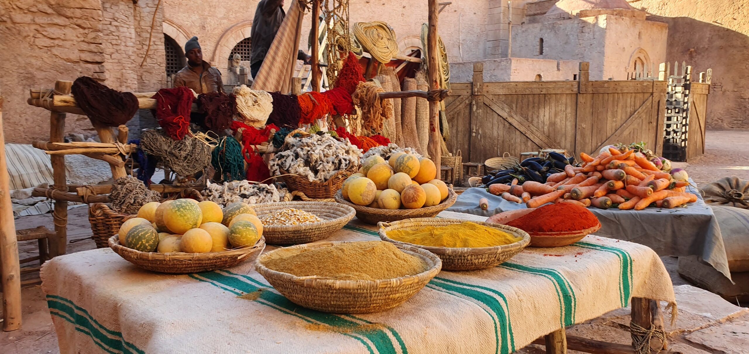 Colorful outdoor market scene featuring fresh spices, melons, and fabrics under natural daylight.