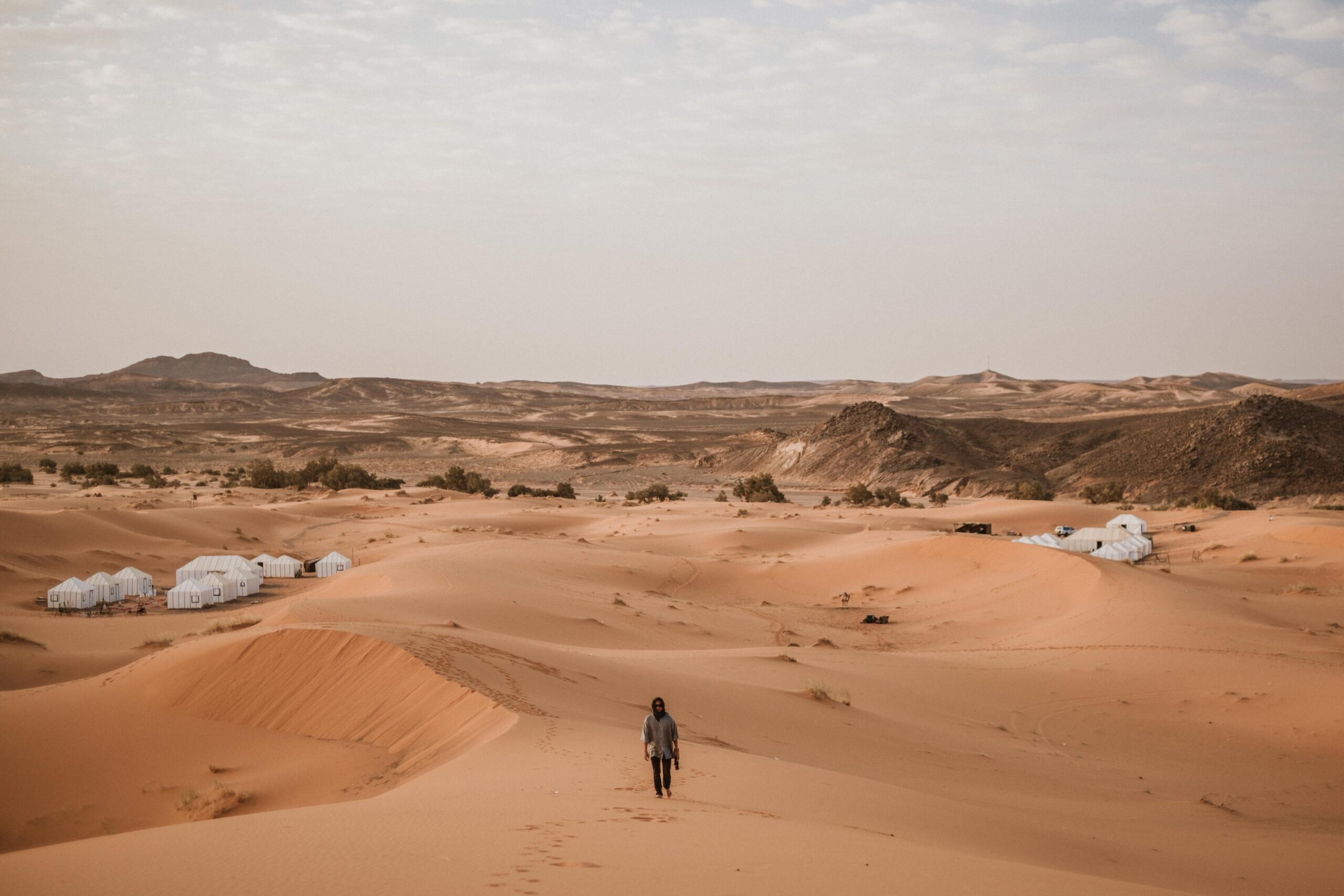A lone traveler walks through the vast sand dunes of Morocco, surrounded by desert landscapes and tents.